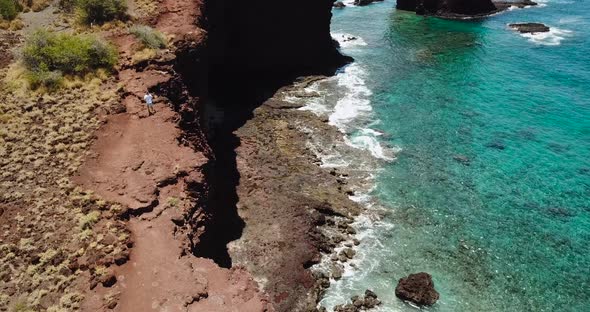 Drone on cliffside beach while man runs on the edge closest to the clear blue water.