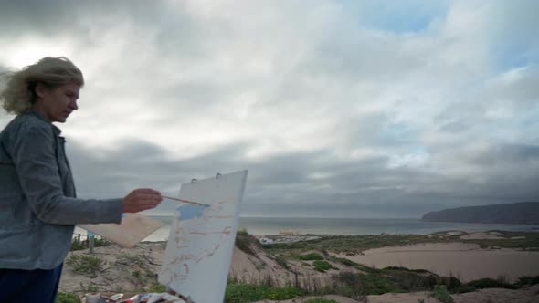 Middleaged European Woman Artist Paints a Picture with Paints Overlooking the Sea
