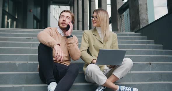 Man and Woman Sitting on Stairs in Urban City Center Working on Laptop, Talking on Mobile phone