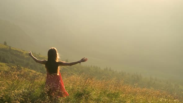 Young Woman in Red Dress Standing on Grassy Field on a Windy Evening in Autumn Mountains Raising Up