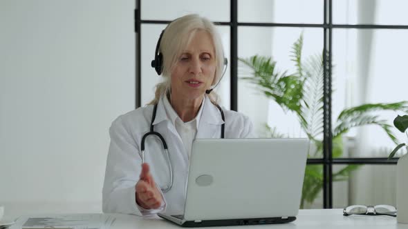 Elderly Female Doctor with a Stethoscope Wearing a Lab Coat and Headset Communicates By Video Call