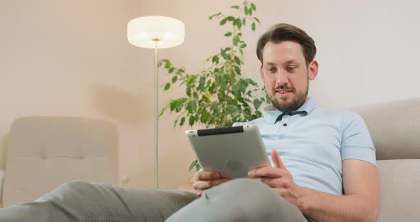 Young Bearded Man is Lying on the Sofa and Holding Tablet in Hands He is Relaxing and Scrolling the