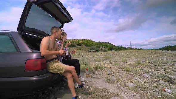 Young happy couple sits in trunk of car and drinks tea