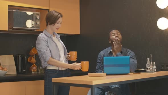 Young Smiling Caucasian Woman Giving Cup of Tea To Busy African American Man Working Online in