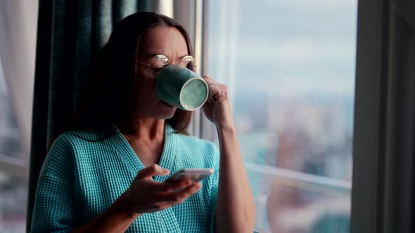 a Beautiful European Woman Drinks Coffee at Home Using Her Smartphone Enjoying a Relaxed Morning