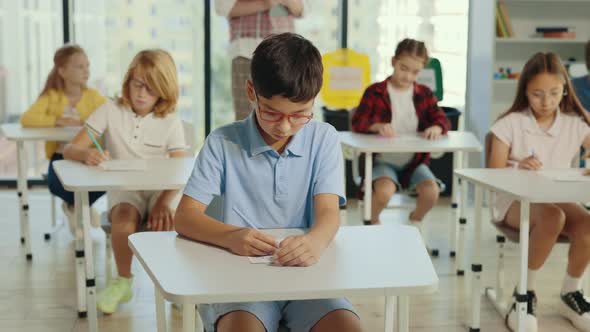 Asian Boy Pupil Wearing Glasses Sits at a First Desk in the Classroom and Fills Out Tests Thinks