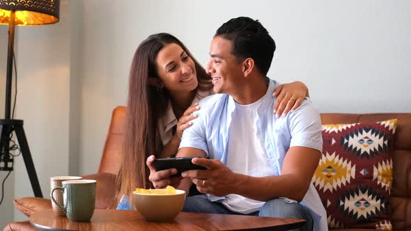 Young happy couple use smart phone together at home sitting on the couch - cheerful girl and boy