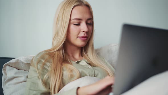 Happy blond woman working on laptop in bed