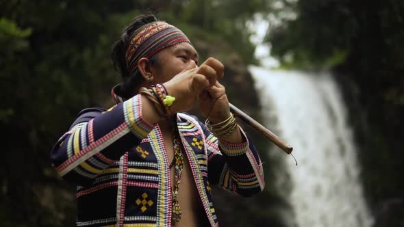 orbit shot of Indigenous person playing Kubing tribal instrument, behind is a waterfall while standi