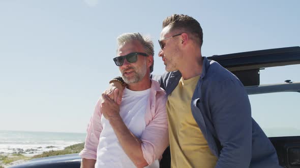 Happy caucasian gay male couple wearing sunglasses embracing by car on sunny day at the beach