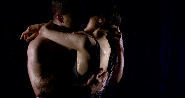 A Muscular Man Passionately Hugs and Kisses a Woman in the Pouring Rain on a Black Background Close