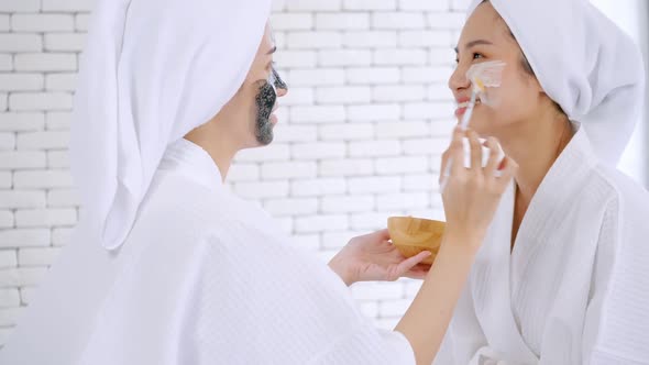 Beautiful young woman in white bathrobe applying a revitalizing white mask onto her friend's face.