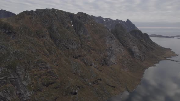 Aerial drone view flying parallel to a steep, rocky mountain wall in cloudy Lofoten, Norway