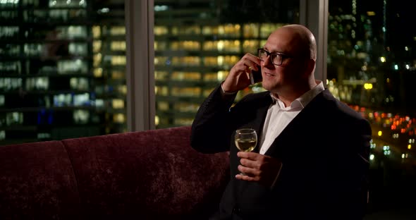 Adult Bald Man Is Talking By Phone and Drinking White Wine, Sitting in His Flat at Night