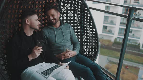 Young Handsome Gay Couple Relaxing on the Swing on the Balcony with Beer and Browsing Social Media