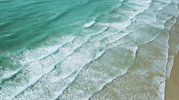 Wide aerial-shot of crystal clear waves crashing onto a sandy beach.