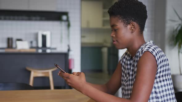 African american businesswoman wearing face mask sitting in workplace kitchen using smartphone