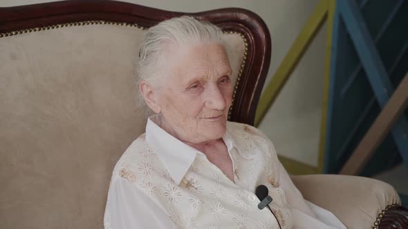 Portrait of Cute Grandmother Sitting on Sofa in Room and Talking at Camera