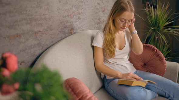 Restless Woman in Glasses Reads Book Sitting on Sofa