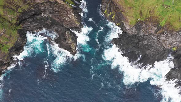 Aerial View of Waves Break on Rocks of Faroe Islands Cliffs in a Blue Ocean