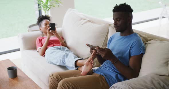 Video of happy african american couple relaxing on sofa with tablet and smartphone