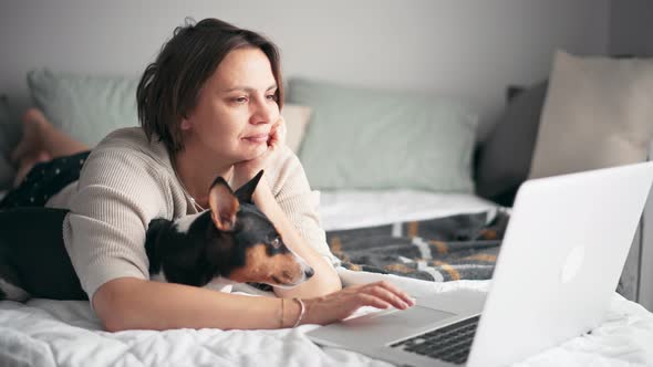 A Middleaged Woman Is Working on Her Laptop While Lying on the Bed with Her Dog
