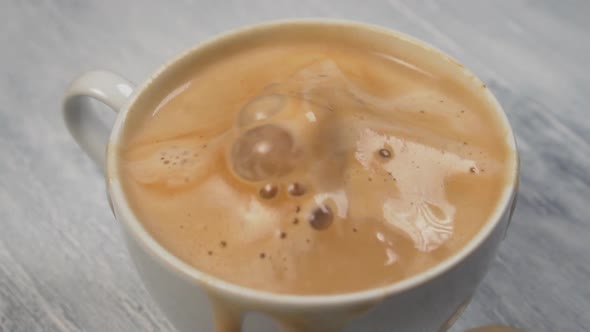 Ice cube falls into white ceramic cup of frothy coffee 