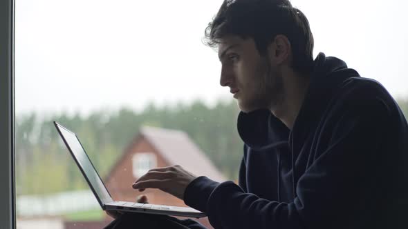 A Young Man is Typing on a Laptop Sitting at the Window Against the Backdrop of a Picturesque Rural