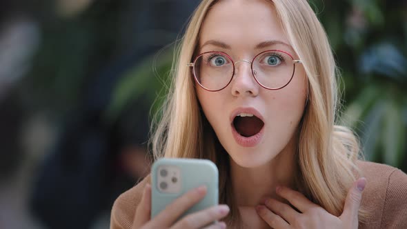 Portrait Caucasian Girl Blonde Successful Lucky Woman in Glasses Looks at Screen of Mobile Phone