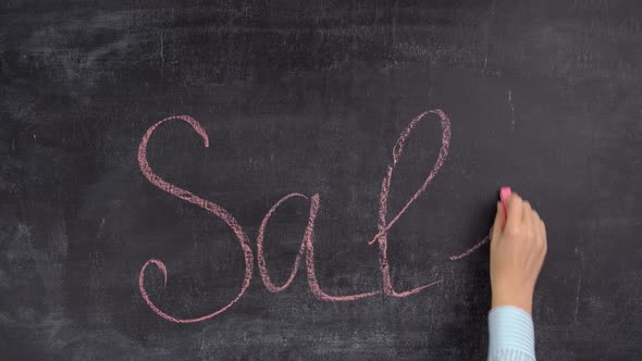 A woman's hand writes the word SALE in red chalk on a chalkboard