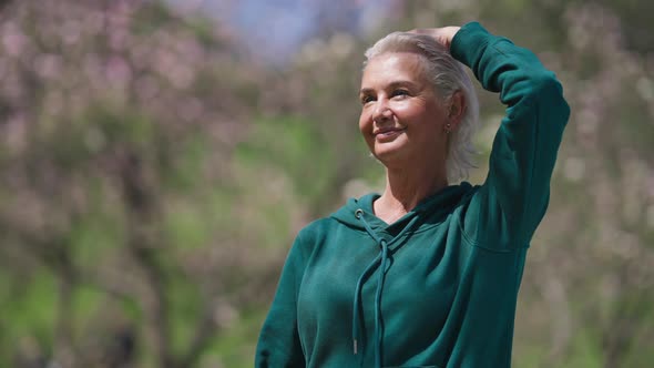 Cheerful Senior Woman Stretching Neck Muscles Training in Sunny Summer Park