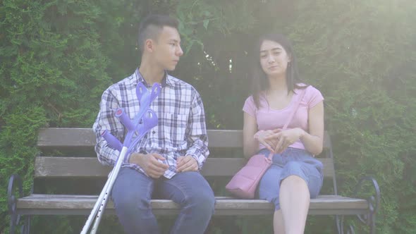 Young Positive Asian Man with Broken Leg on Crutches Communicates with Young Asian Woman Sitting on