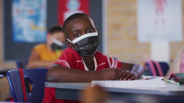 Portrait of african american schoolboy wearing face mask, sitting in classroom looking at camera