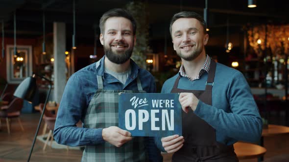 Portrait of Men Business Partners Holding We Are Open Sign Standing in Modern Cafe