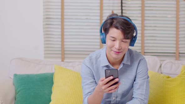 Asian handsome man sit on couch wearing headphone listen music and using mobile