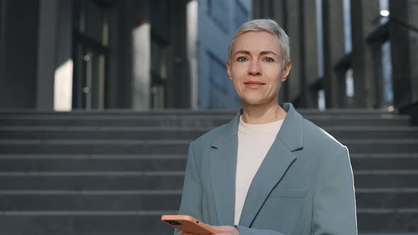 Beautiful Middle Aged Woman in Formal Wear Using Modern Smartphone Looking at Camera Standing at