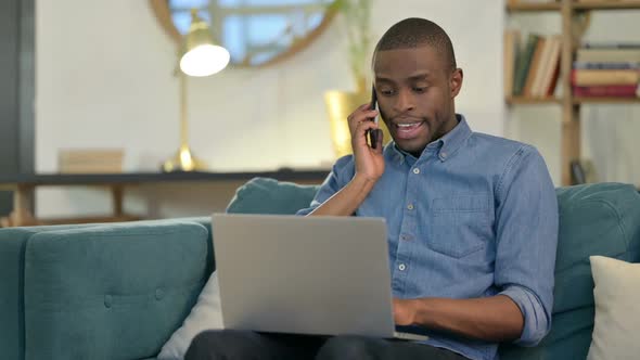 Young African Man with Laptop Talking on Smartphone on Sofa