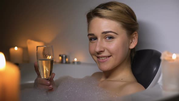 Girl in Bath With Foam and Candles Holding Glass of Champagne, Smiling at Camera