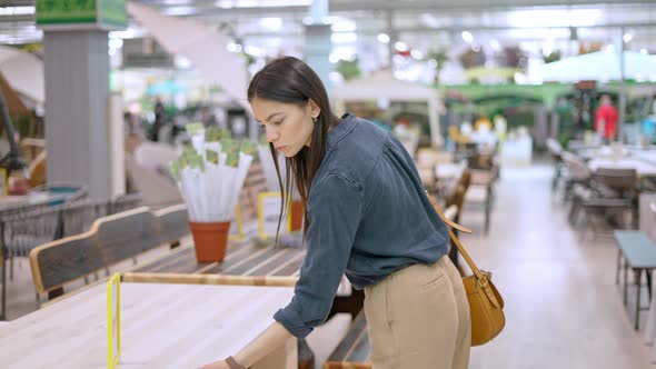 Young Focused Woman Looking at Price of Wooden Table While Choosing Furniture for Garden
