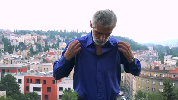 Old Senior Man Adjusts Clothes and Smiles To Camera - City (Buildings) in Background