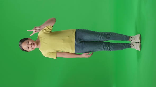 Full Body Of Smiling Young Asian Kid Girl Showing Gesture Peace While Standing On Green Screen alt
