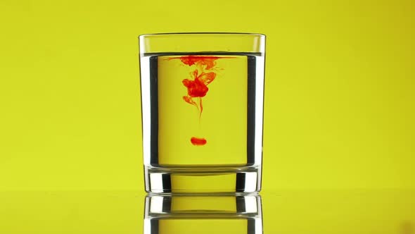 Two Red Drops of Ink Dripping Into Clear Water in Glass Standing on Yellow Background in Slowmotion