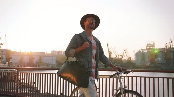 A Young Stylish Hipster with Hat and Glasses Walking with His Bicycle on Sea Port Background During