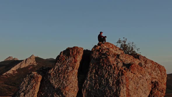 A Man Sits On Top Of A Mountain Which Is Illuminated By The Last Rays Of The Sunset