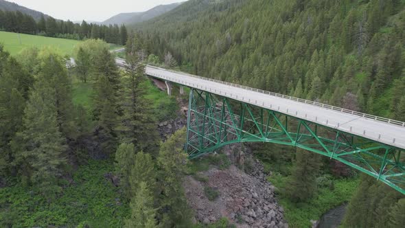 Drone is rising above a steel bridge at the mouth of a pine covered canyon.  The road can be followe