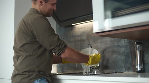 Young Man in Gloves Washing Dishes While Standing in Kitchen.