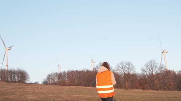 Female Engineer in Orange Vesta Goes to Wind Turbines with a Tablet to Checks Their Operation