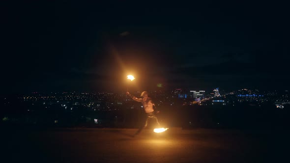 Young Blond Male Dances with Two Burning Torches in the Middle of the Night with City Skyline in