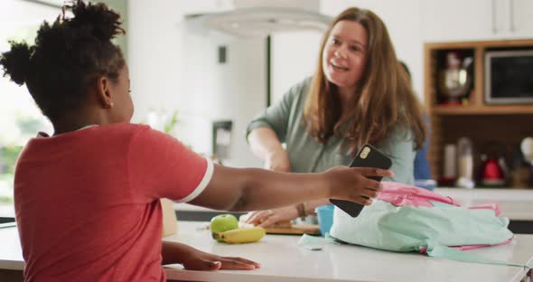 Happy caucasian woman and her african american daughter using smartphone in kitchen