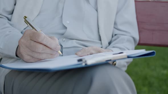 Close View of Old Male Hands Writing on Papers Lying on Knees on Park Bench